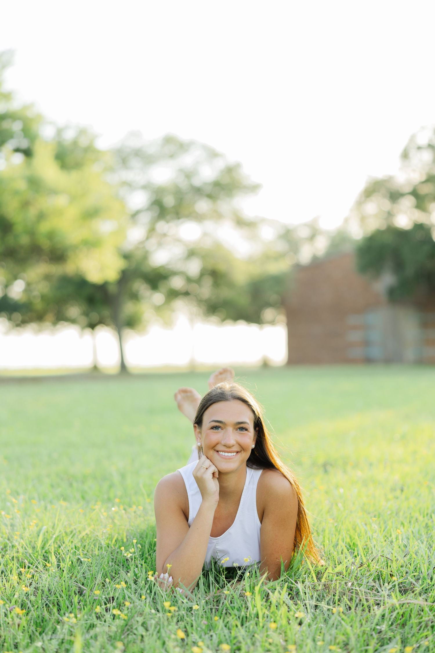 Smiling young woman lying in green grass field with trees and building in background.