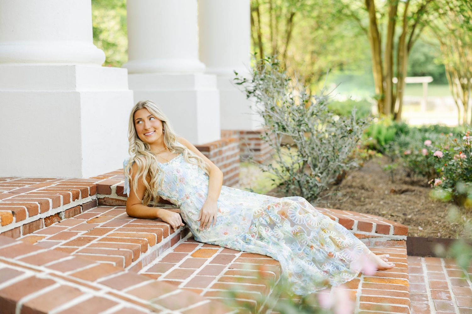 Young woman in floral maxi dress reclines on brick steps beside white columns in a garden.