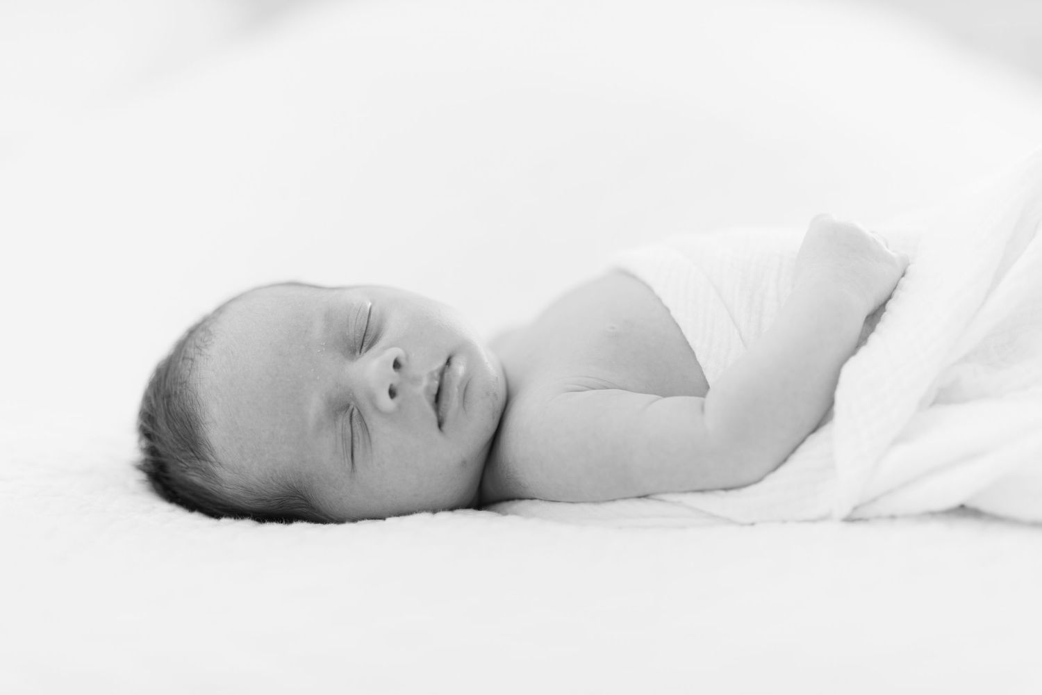 Peaceful newborn baby wrapped in white swaddle sleeping on a white surface, black and white photo.