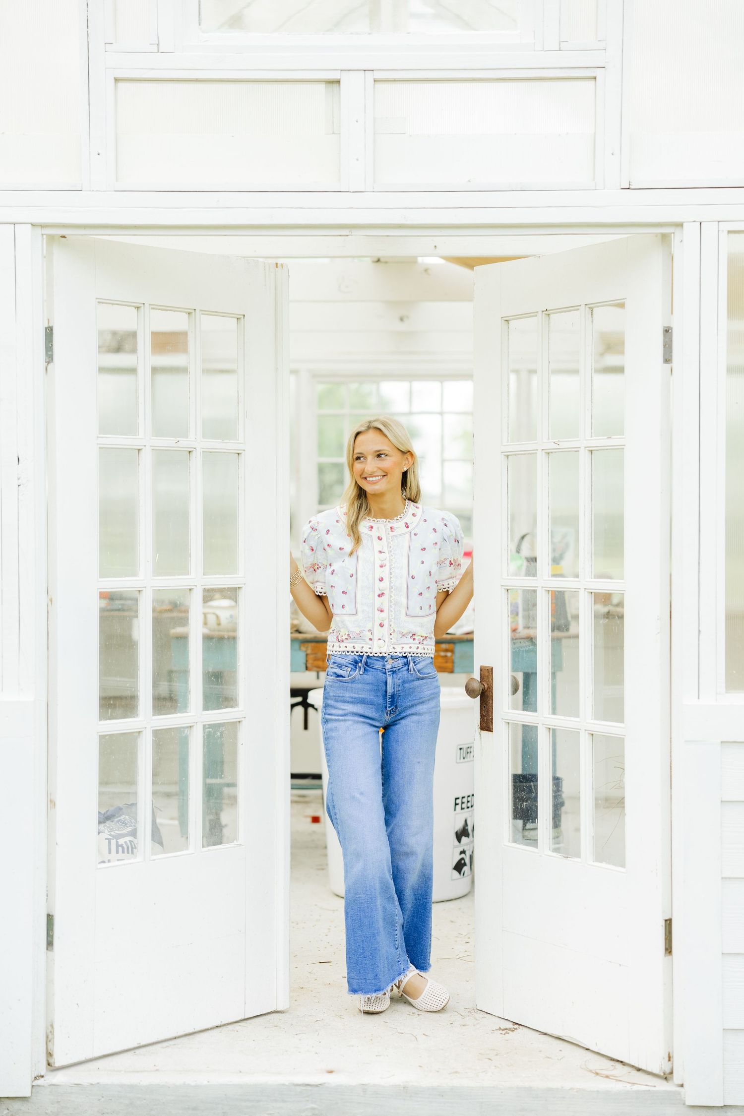 Blonde woman in floral blouse and wide-leg jeans standing in open white French doorway.