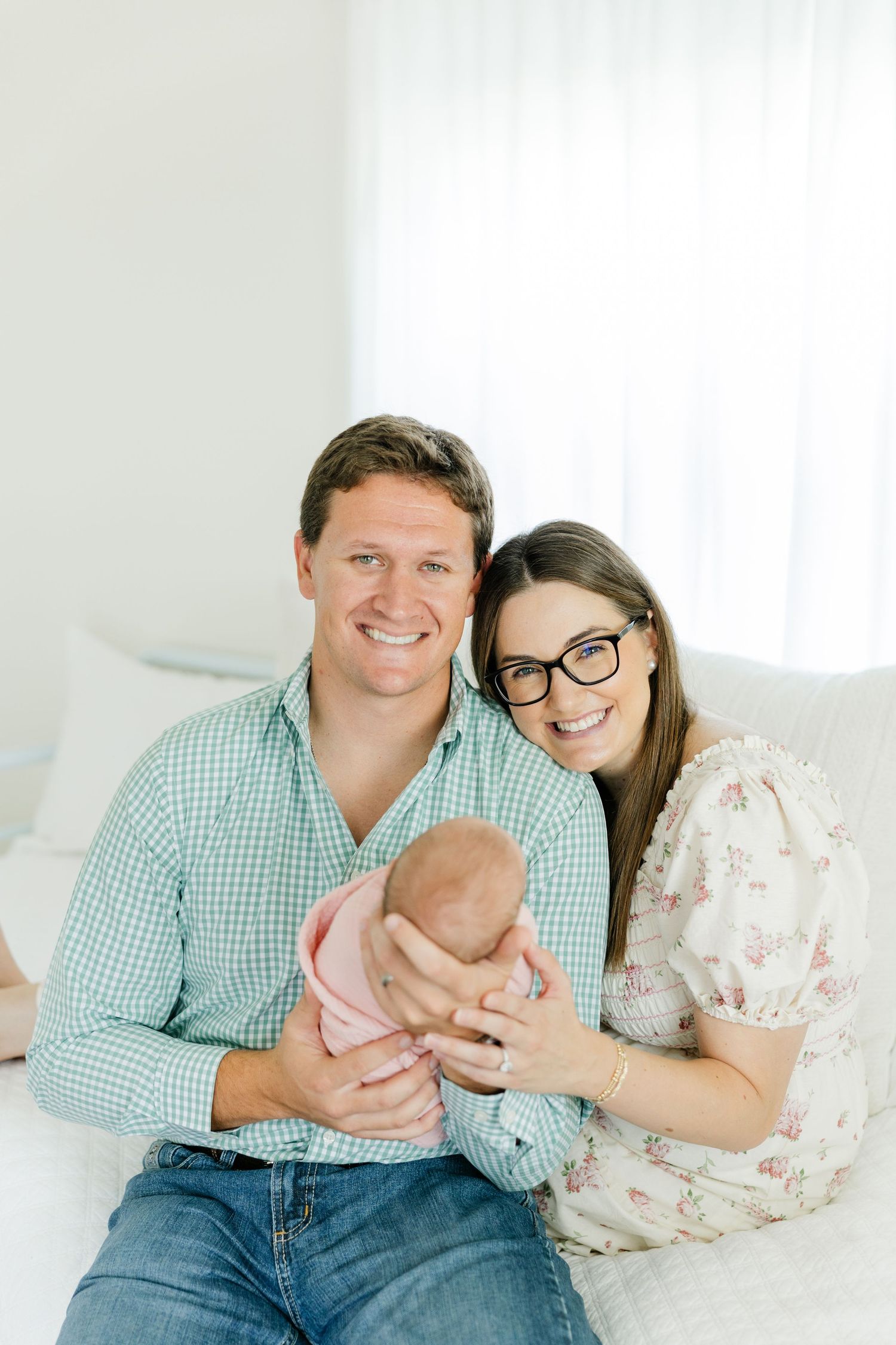 Smiling parents sitting together holding their pink-swaddled newborn baby for a family portrait.