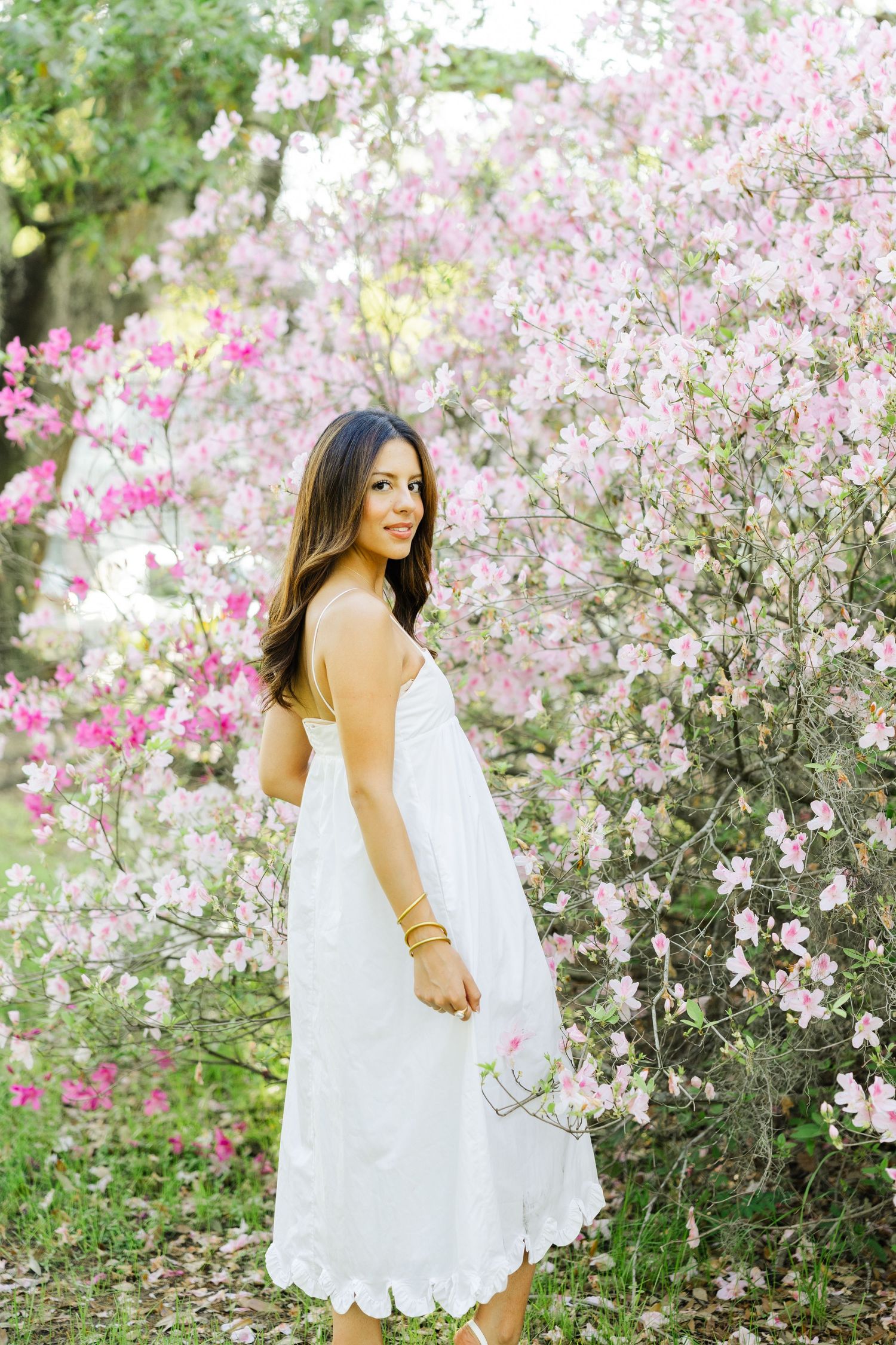 Young woman in white eyelet dress poses among blooming pink azalea bushes in a garden.