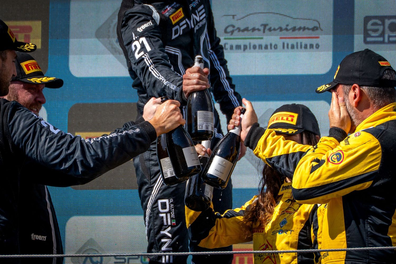 Drivers celebrate on the podium, clinking champagne bottles together in a toast.