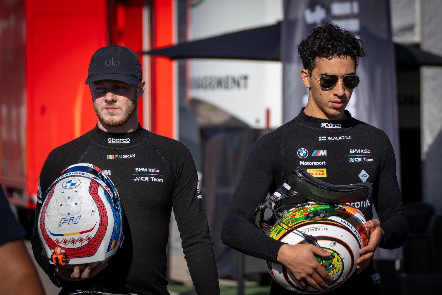 Two racing drivers standing together in the pit lane, holding their helmets before a race.