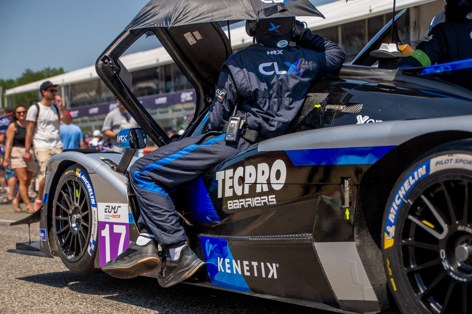 A mechanic in a blue and black suit sitting inside the open gullwing cockpit of a number 17 CLX Motorsport prototype car in a busy grid.