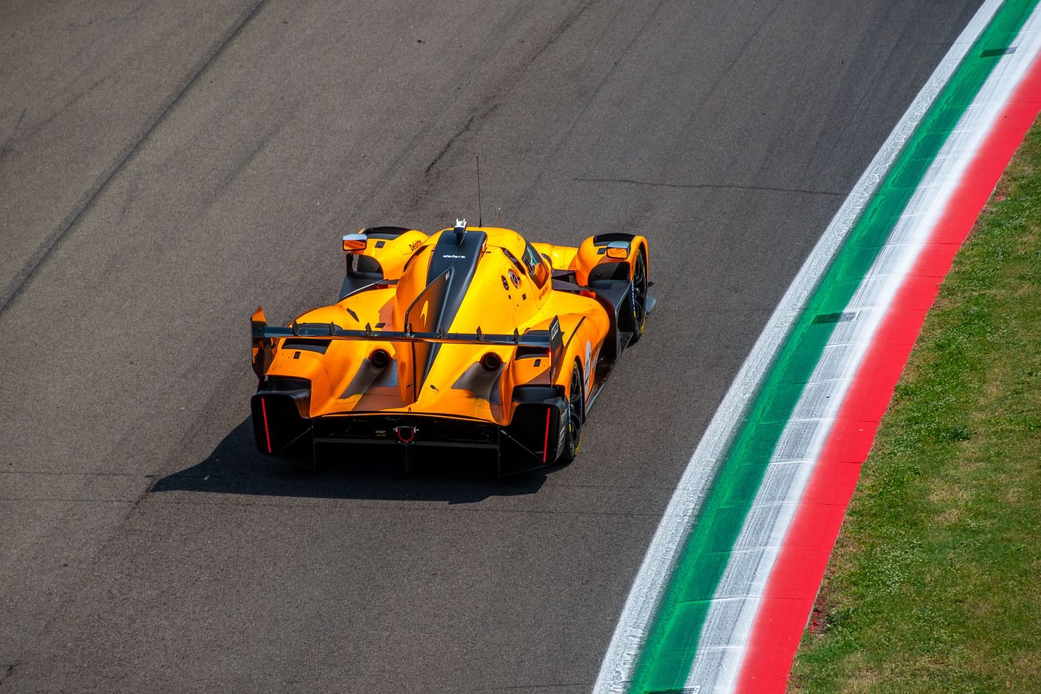 High-angle rear view of the orange and black McLaren Hypercar driving alongside the iconic tricolor curbs at Imola.