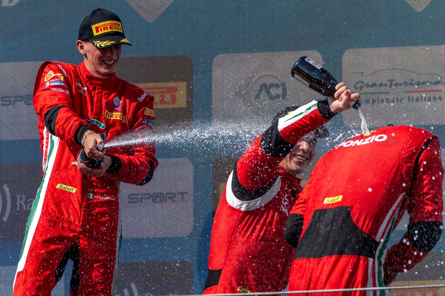 Two Ferrari drivers celebrate on the podium as champagne sprays around them.