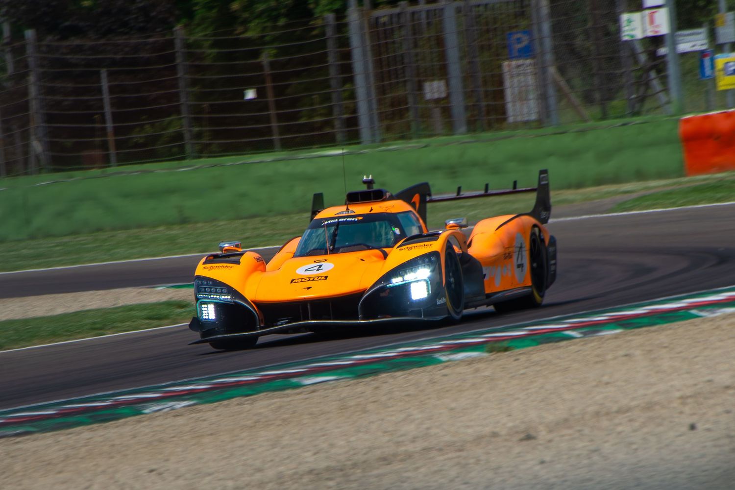 Front three-quarter view of the orange racing car with bright headlights driving through a corner on the racetrack.