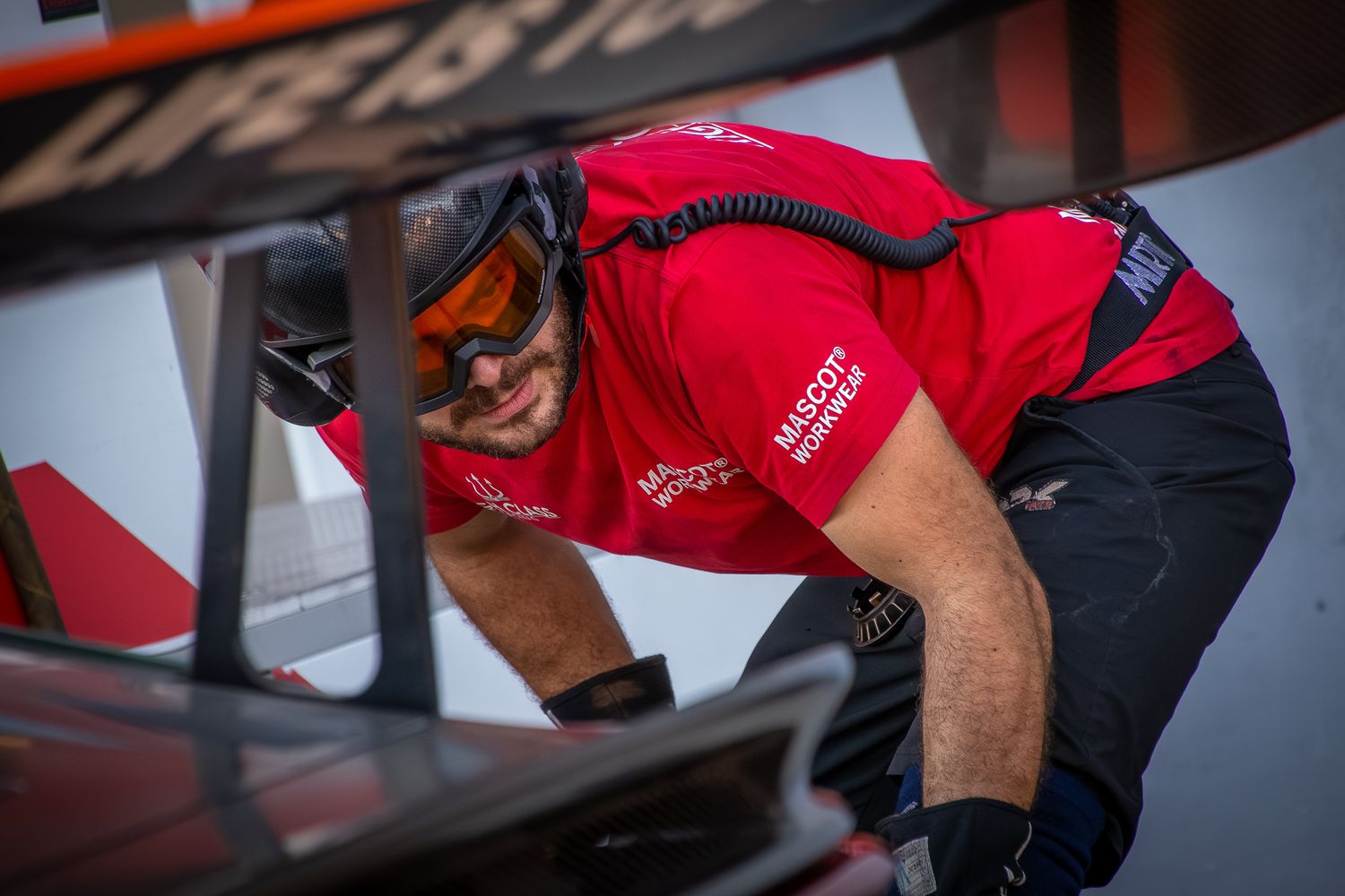 Race team crew member in a red uniform working on the rear wing spoiler of a sports car at the racetrack.