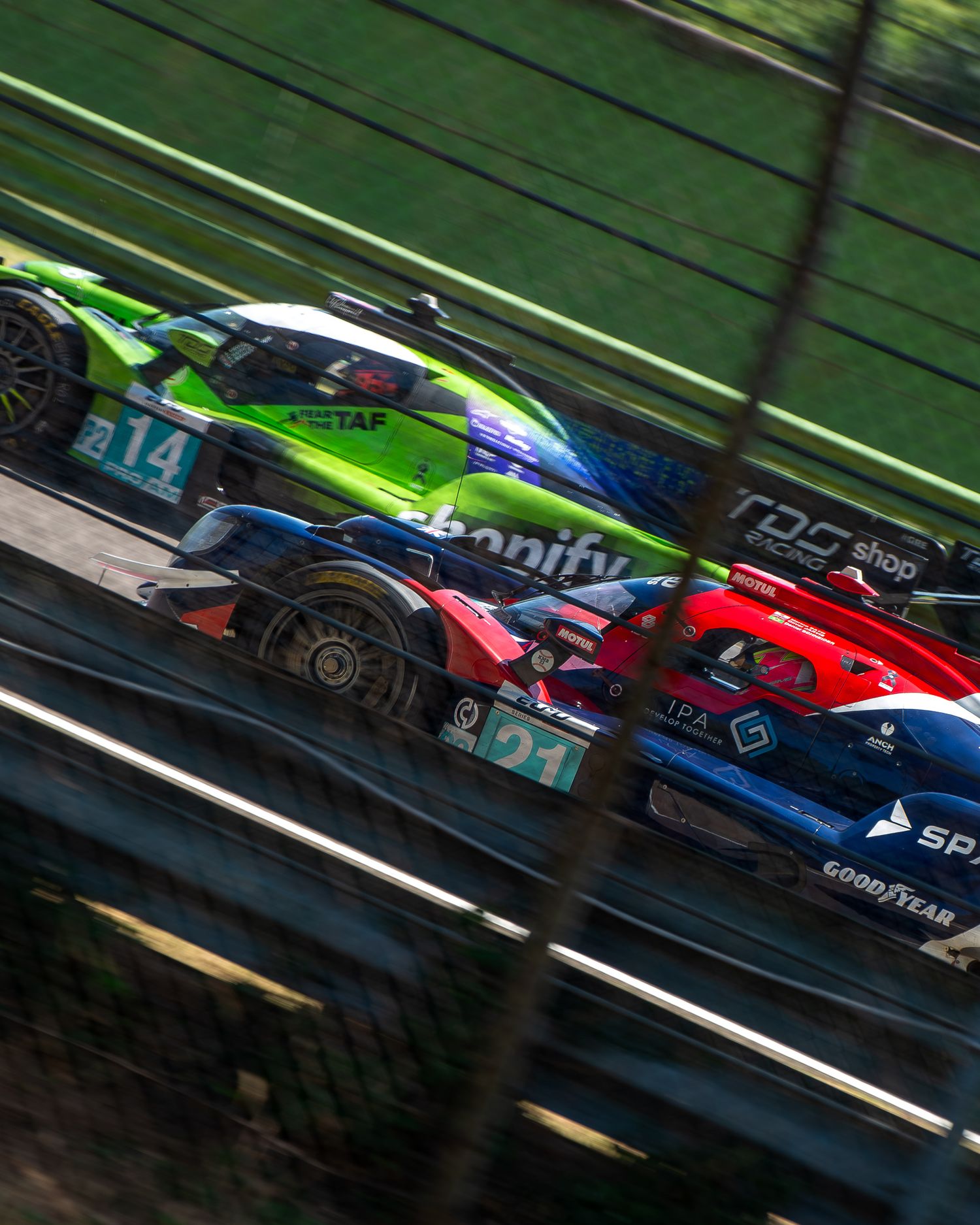 Dramatic Dutch angle shot of a green and a red racing car speeding side-by-side past the track safety barrier.