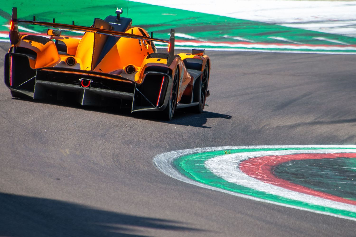 Rear three-quarter view of an orange and black endurance racing car cutting over the painted curbs of a sharp turn.