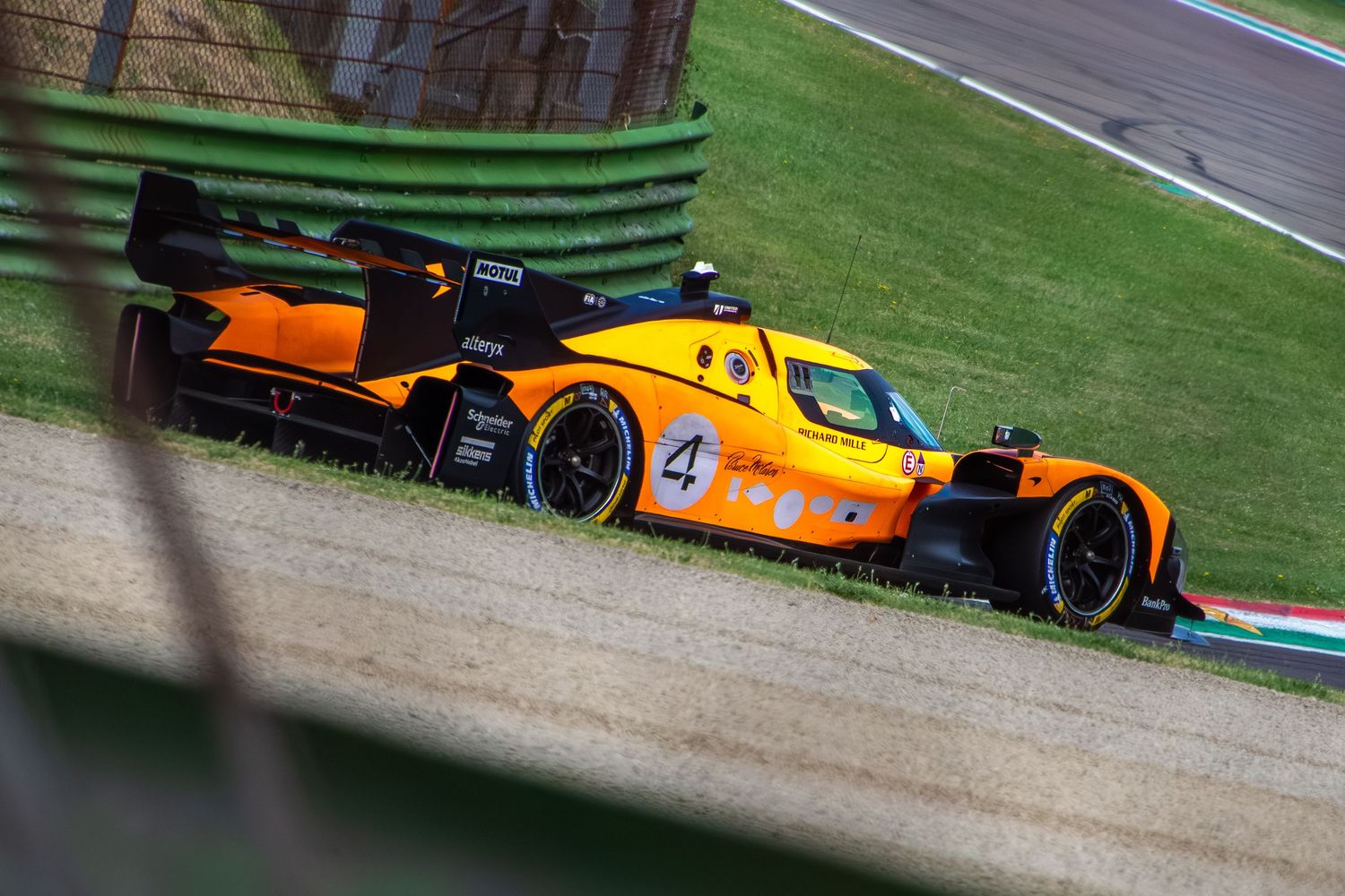 High-angle shot of the orange number 4 prototype racing car speeding alongside the green trackside grass hill.
