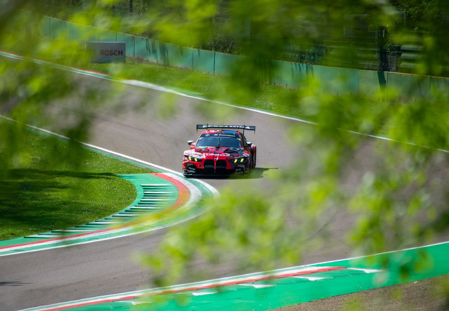 A red and white BMW M4 GT3 racing on a track, framed by blurry green foliage in the foreground.
