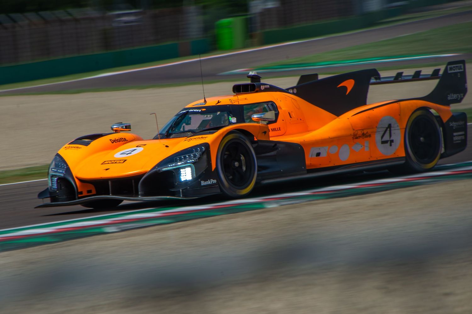 Low-angle panning shot of the orange and black number 4 race car driving fast down a straight section of the track.
