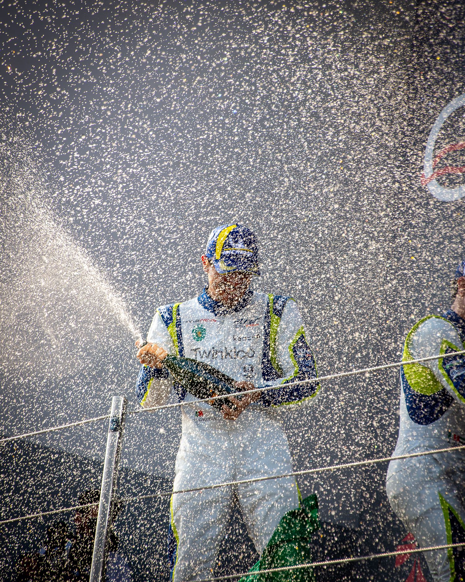 A racing driver celebrating on the podium, spraying champagne into the air with a dramatic backlit splash effect.