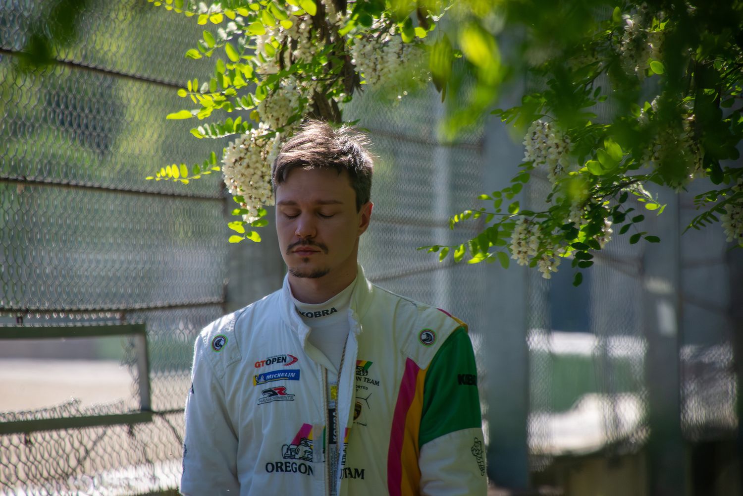 A race car driver standing thoughtfully behind a wire fence under hanging green leaves and flowers.