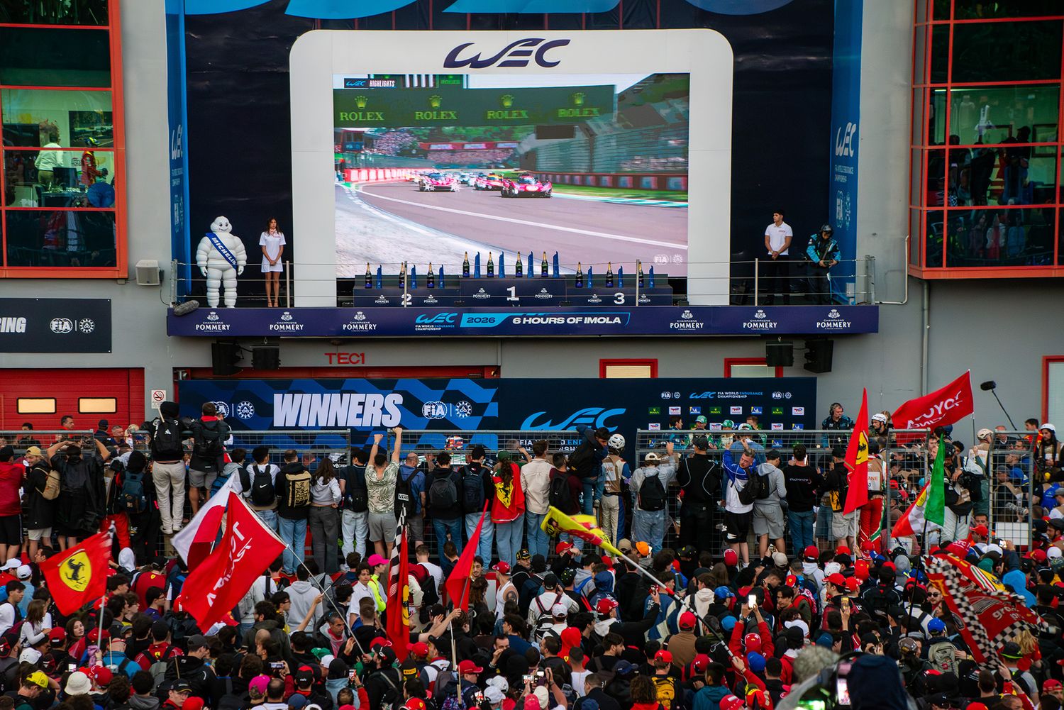Large crowd waving flags in front of the WEC winners podium screen celebrating a race victory.