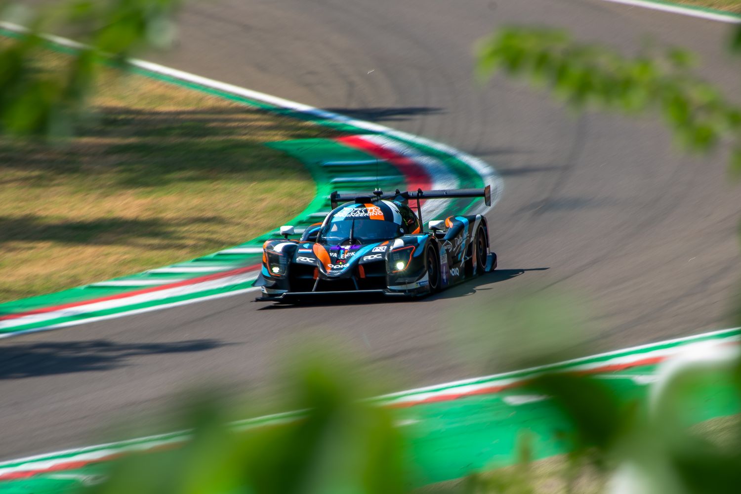 Dark prototype racing car speeding through an S-curve on a racetrack, framed by green leaves in the foreground.