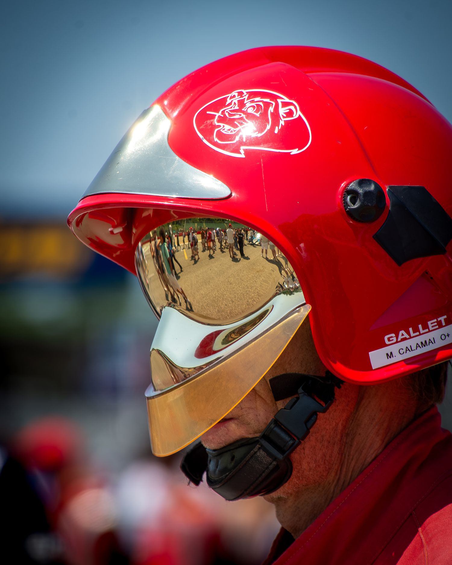 Close-up profile portrait of a track marshal wearing a glossy red helmet with a highly reflective gold mirrored visor reflecting the grid.