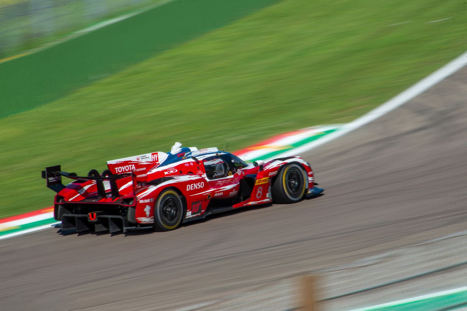 High-angle panning shot of a red and white Toyota TR010 Hypercar speeding past a tricolor curb.