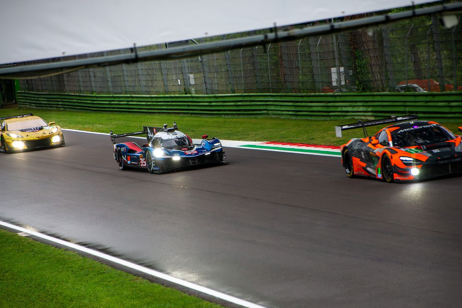 Three endurance race cars driving through wet conditions on the track at Imola circuit.