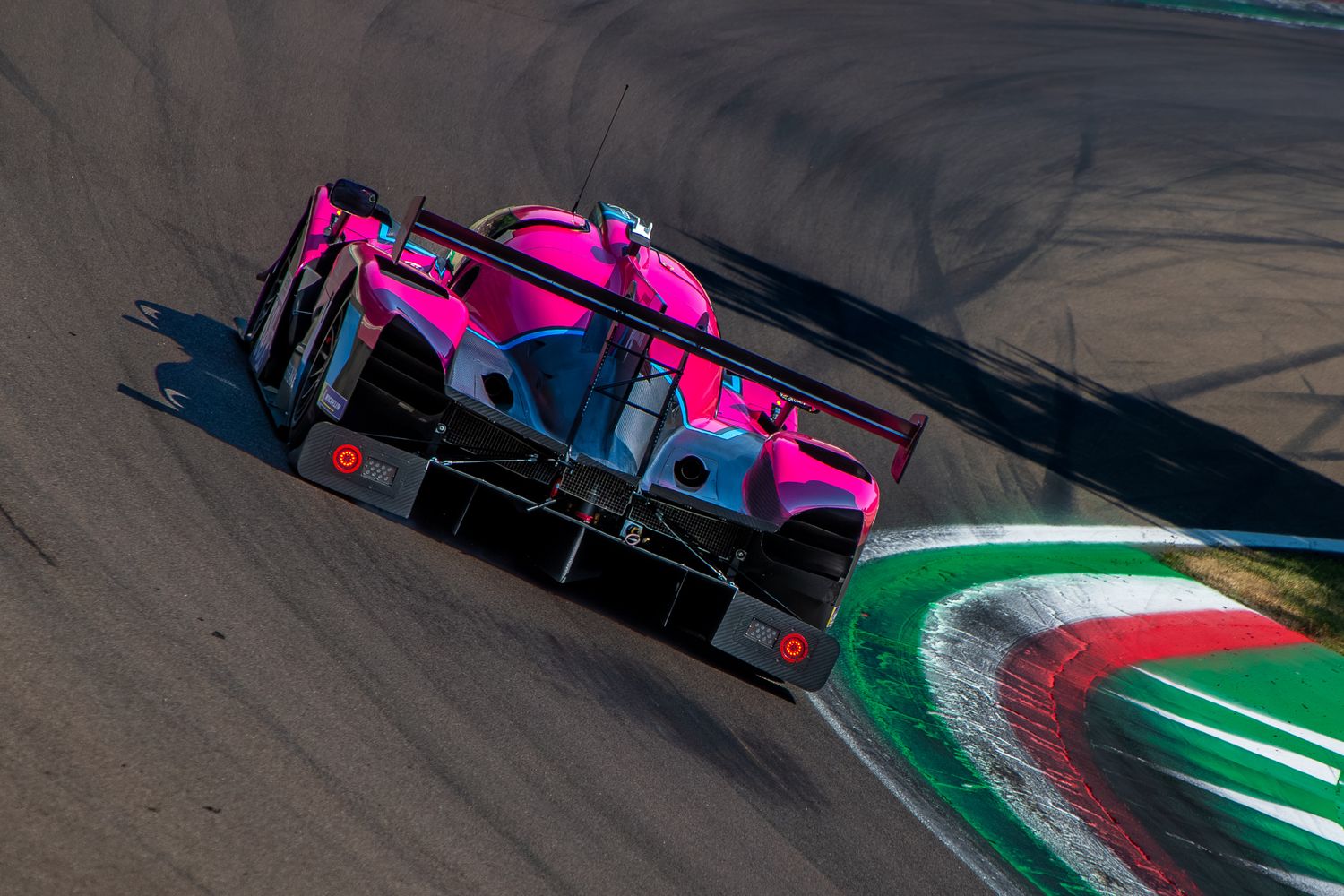 High-angle rear view of a pink and black endurance racing car taking a sharp turn over the track curbs.
