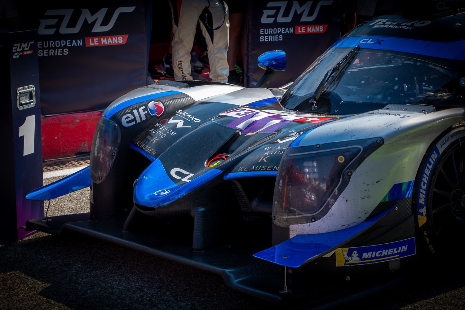 Close-up of a blue and black ELMS prototype racing car under teh podium with the winner board.