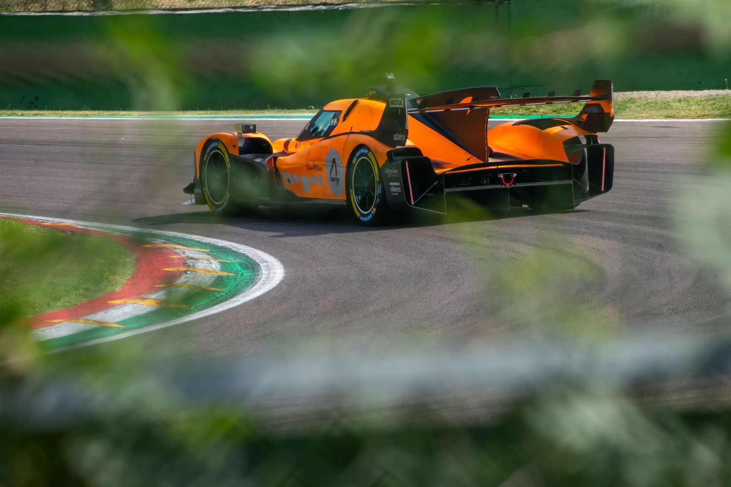 Rear view of the orange endurance race car navigating a corner, framed by a blurred foreground barrier.