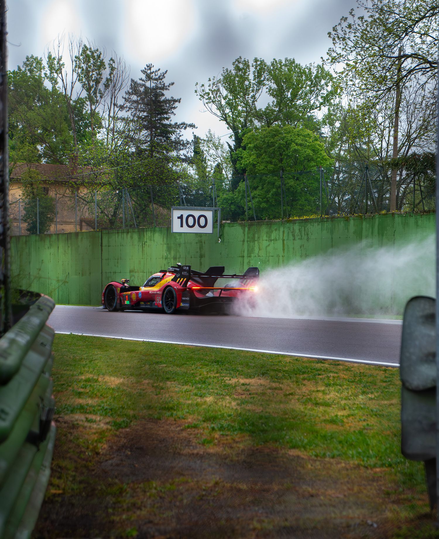 A racing car kicking up water spray on a wet track surrounded by trees at Imola circuit.