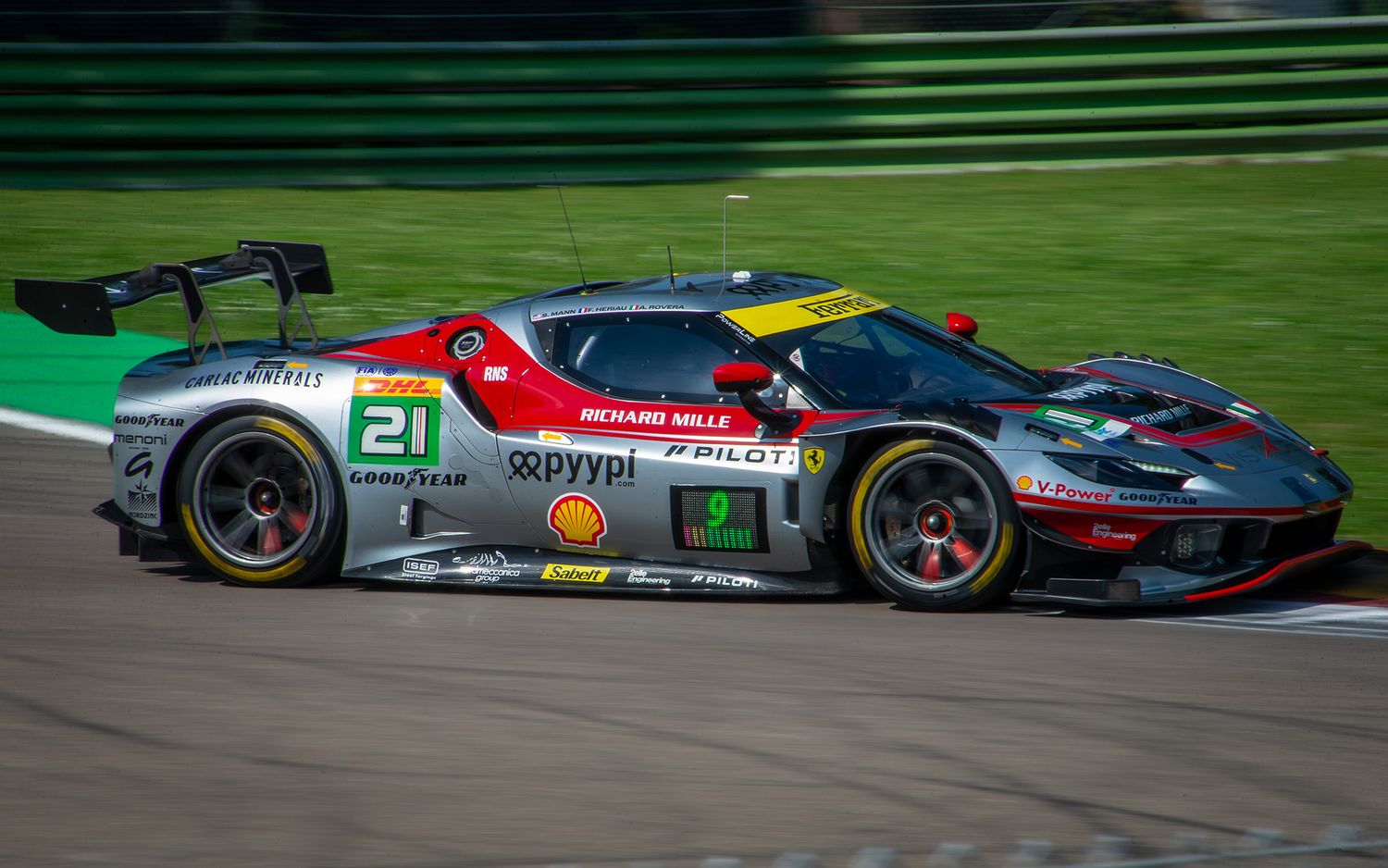 Silver and red Ferrari 296 GT3 racing car driving past the green grass fields at Imola.
