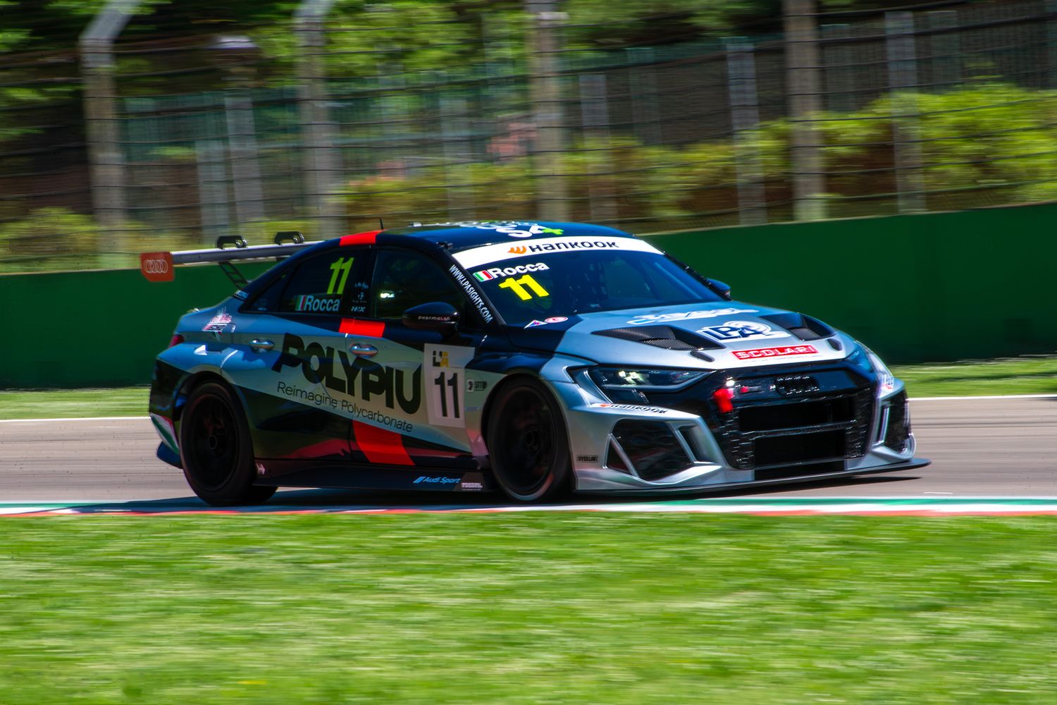 Panning shot of a grey Audi RS3 TCR racing car speeding past the green grass and trees at Imola.