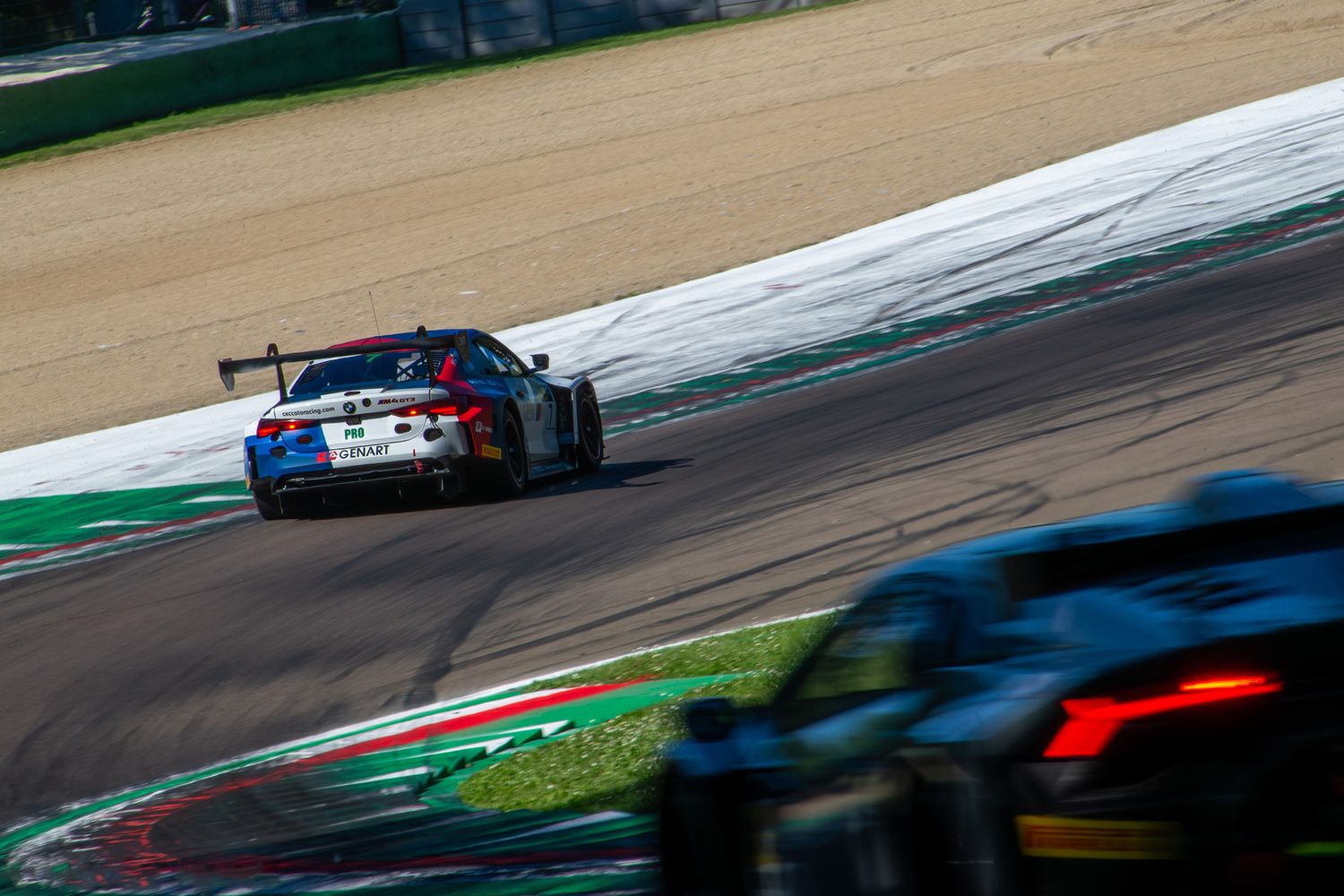 BMW M4 racing car cornering at Imola circuit with another blurred sports car in the close foreground.