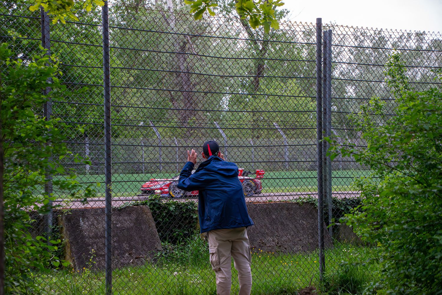 Spectator in a blue jacket watching a red race car zoom past from behind a tall safety fence.