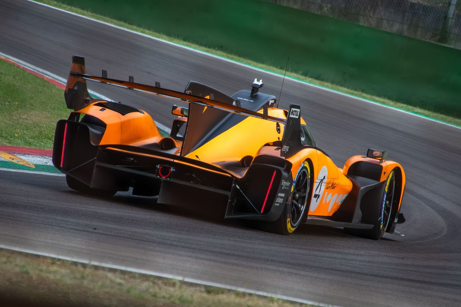 Low-angle rear view of the orange endurance car accelerating out of a corner on the racetrack.