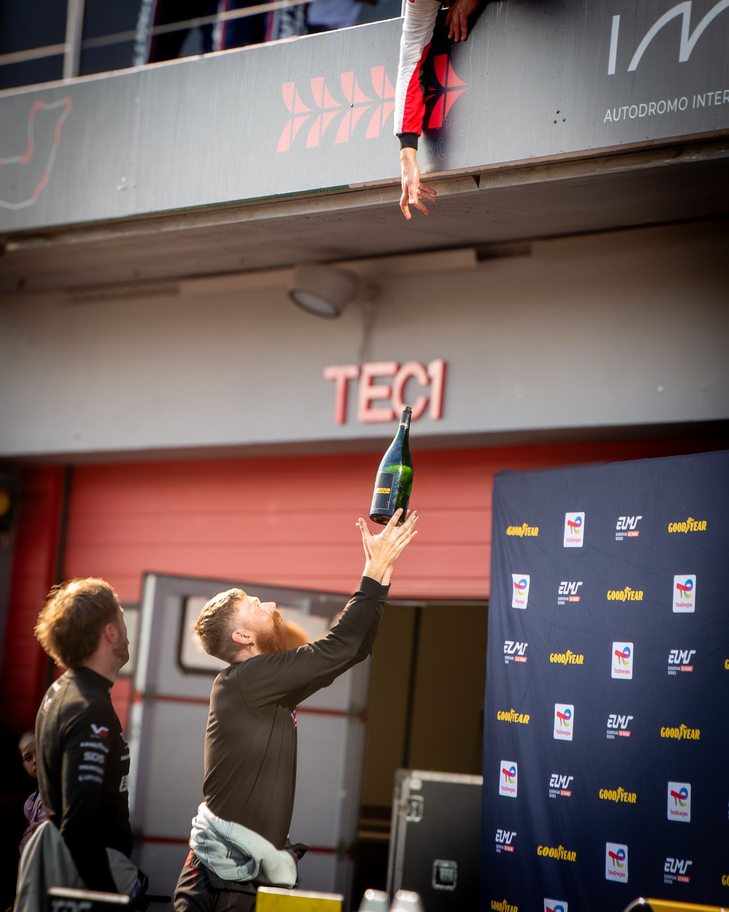 A driver reaching down to hand a champagne bottle to the team member celebrating under the podium balcony above.
