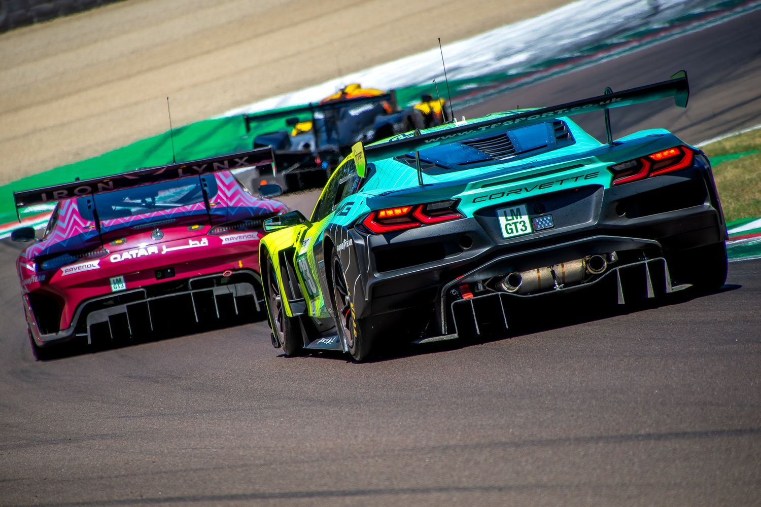 Rear view of two GT3 race cars, a teal and black Corvette and a purple Mercedes, navigating the Acque Minerali turn at the Imola circuit.