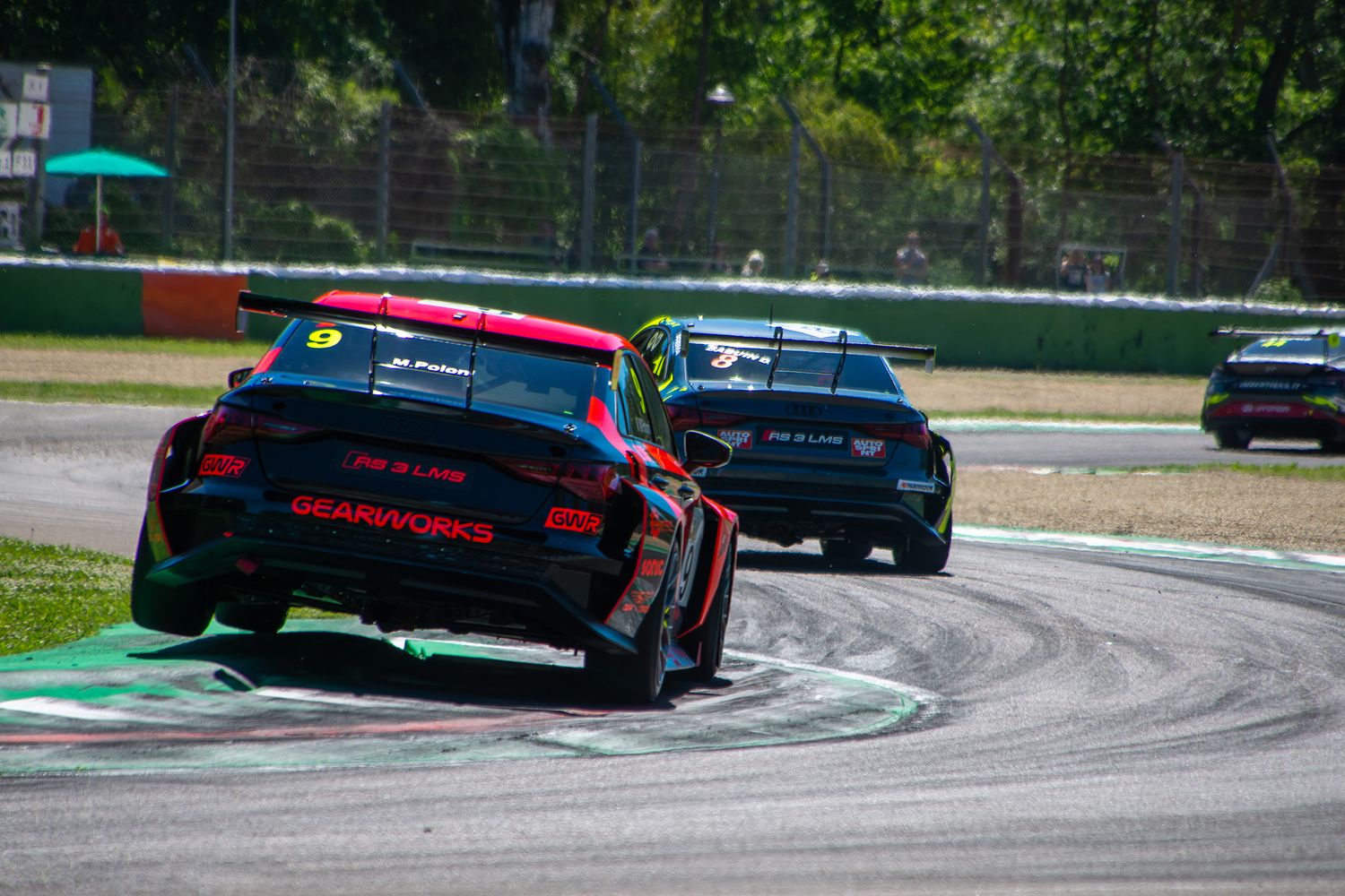 An Audi TCR race car lifting its wheels while driving aggressively over a curb at Imola circuit.
