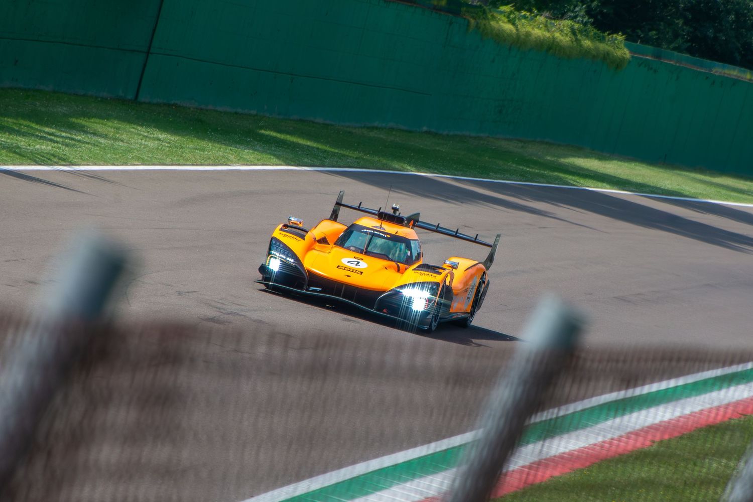 Front view of a bright orange prototype racing car cornering on a racetrack, framed through a trackside fence.
