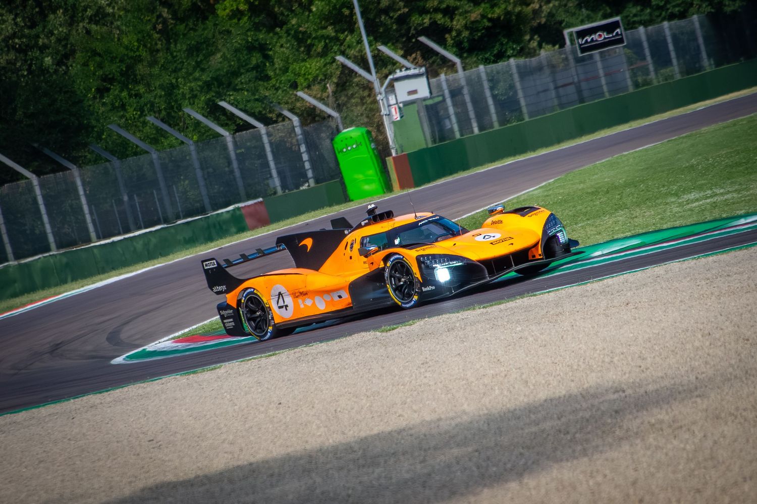 Wide Dutch angle shot of the orange prototype racing car speeding through a curved section of a tree-lined circuit.