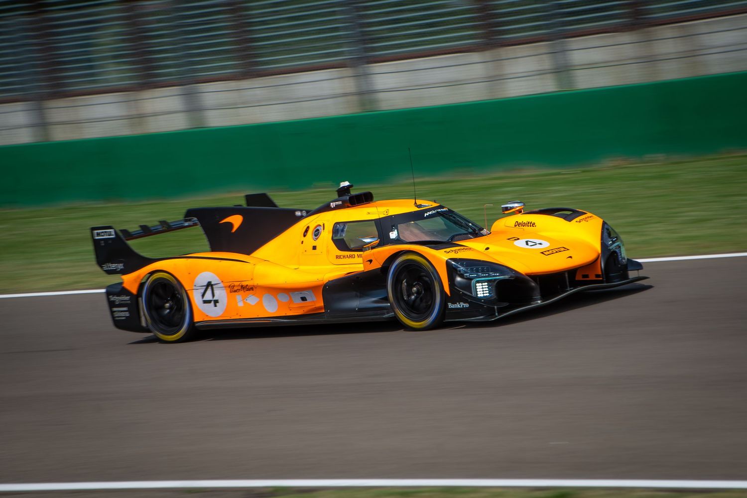 A full side profile shot of the papaya orange and black McLaren Solus GT track car speeding across the dark grey asphalt of a race circuit.