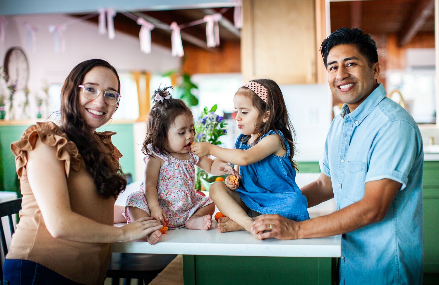 Happy family of four with two young girls smiling together at a table in a colorful indoor setting.