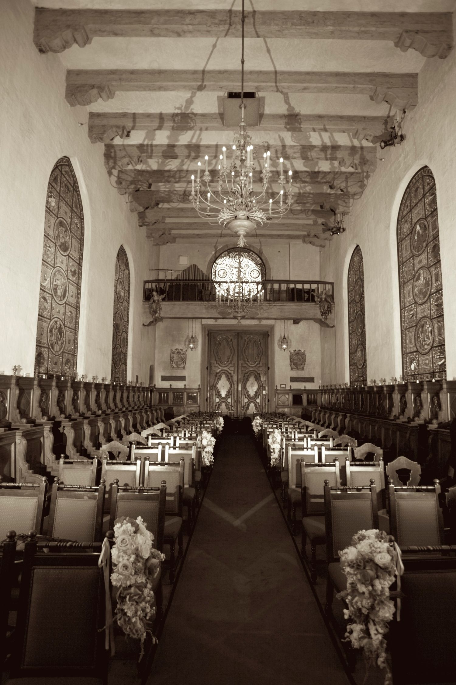 Elegant sepia-toned wedding venue with chandelier, arched walls, floral aisle decor, and ornate wooden chairs leading to altar.