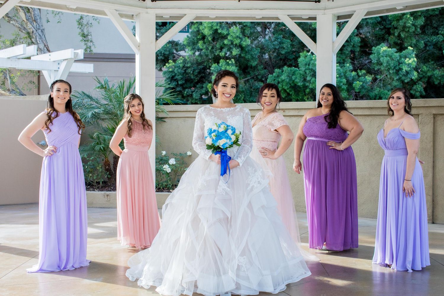 Bride in white ball gown holds blue bouquet surrounded by bridesmaids in lavender and pink dresses at outdoor venue.