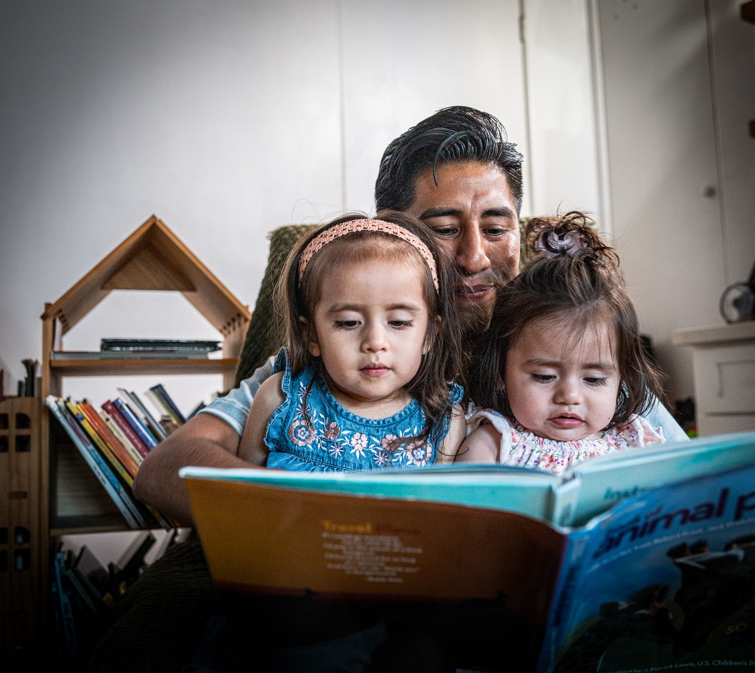 A father reads an illustrated book with his two young daughters at home.