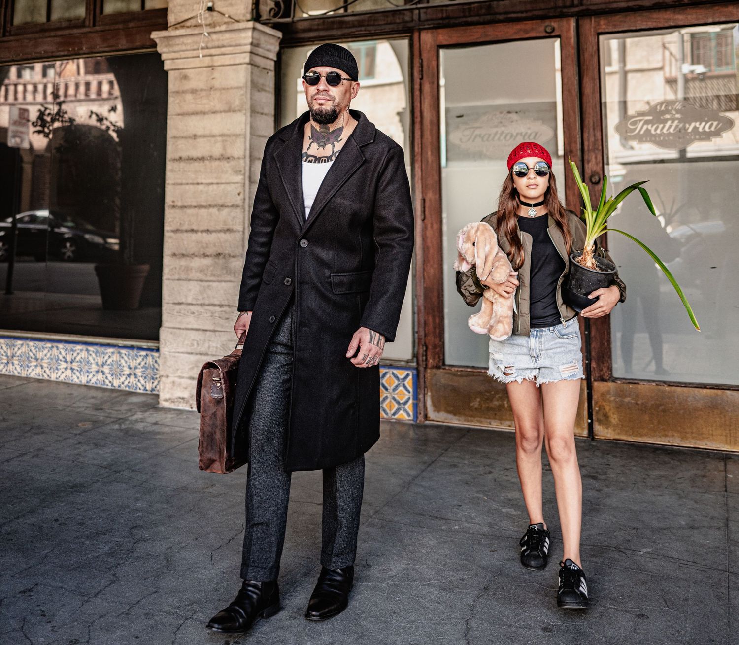 A tall man in a long black coat and a young girl in shorts and a red cap pose with a leather briefcase and potted plant outside a trattoria.
