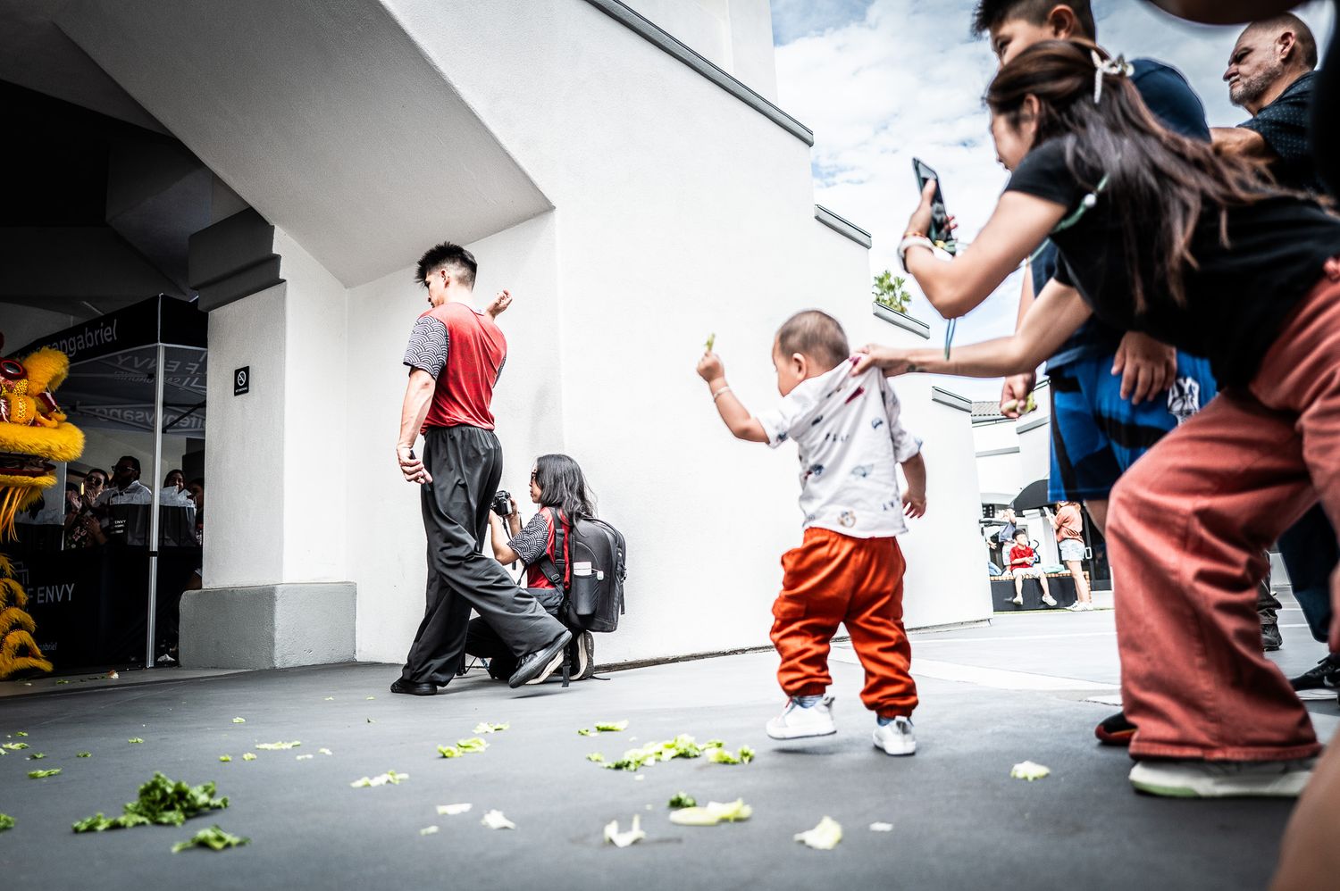 Toddler in orange pants reaches out toward a crowd on a plaza scattered with green petals while onlookers photograph the festive scene.