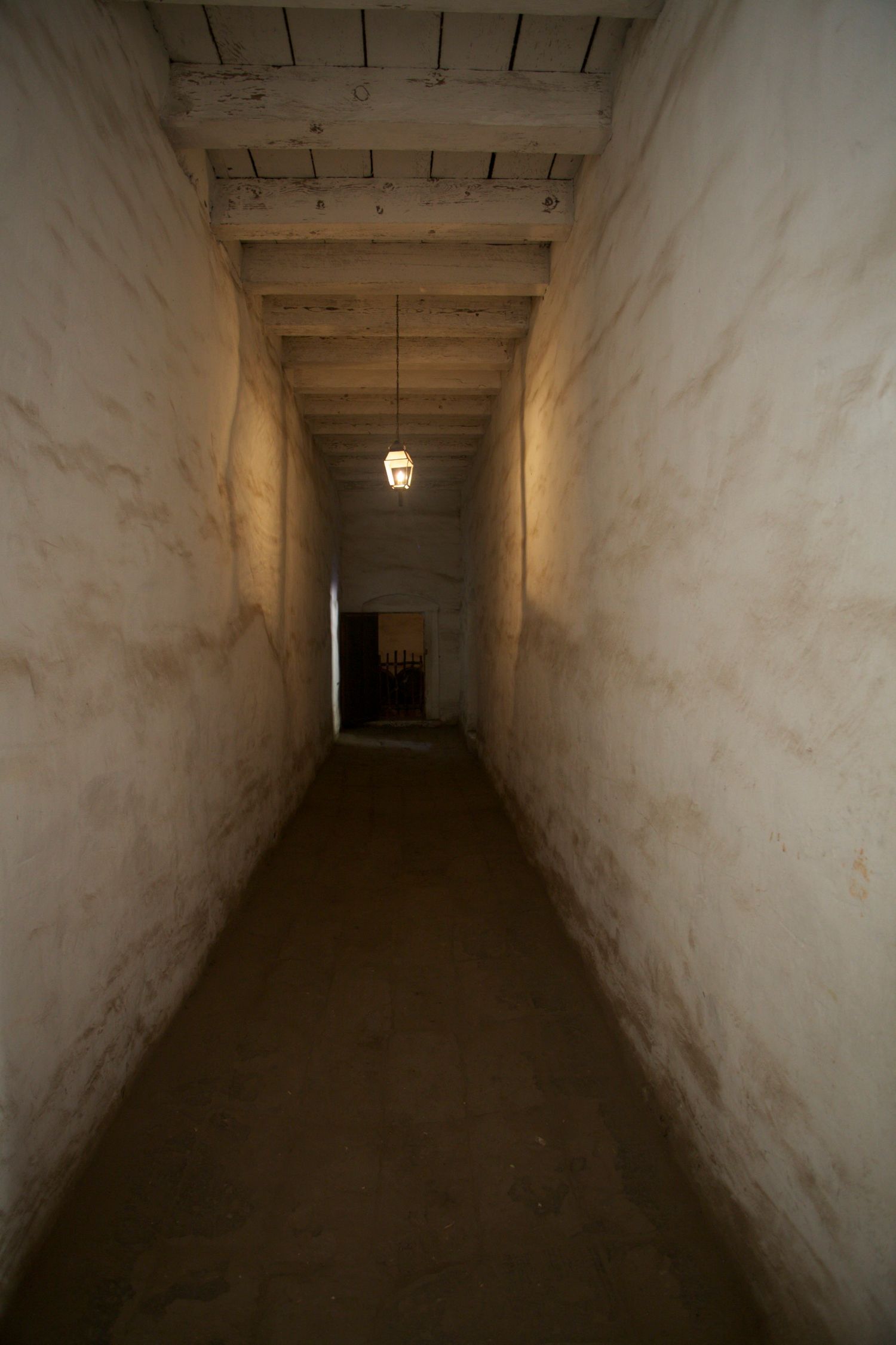 Narrow dimly lit adobe corridor with wooden beam ceiling and hanging lantern leading to a doorway.