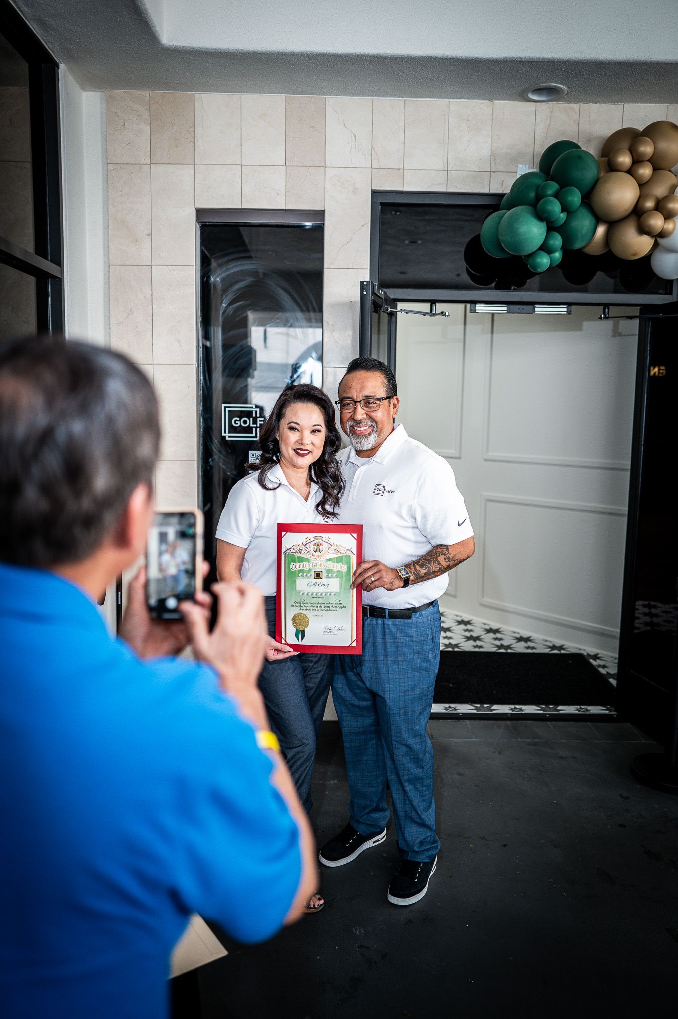 Smiling man and woman in white polos hold a framed certificate while a man in blue photographs them near a green and gold balloon garland.