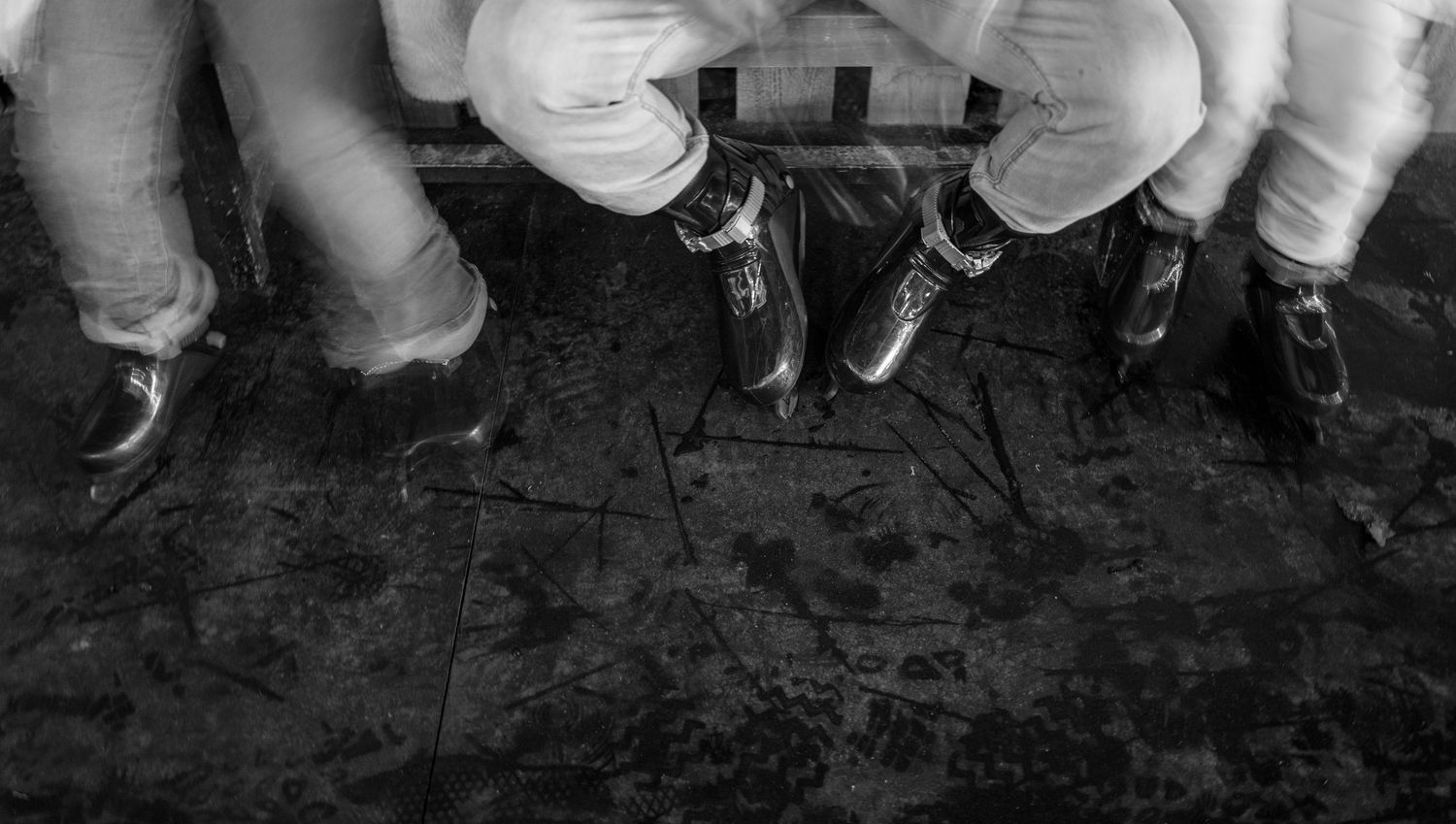 Black and white close-up of three people's dress shoes standing on a worn wooden floor.
