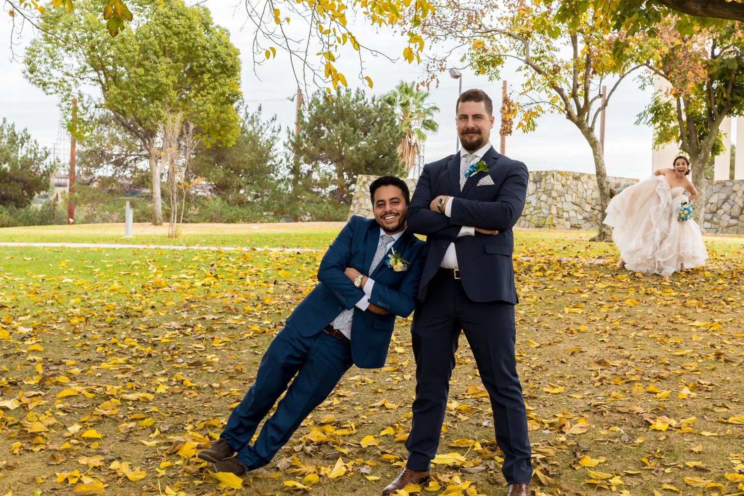 Two grooms pose playfully on autumn leaves at outdoor wedding venue, bride visible in background wearing white gown.